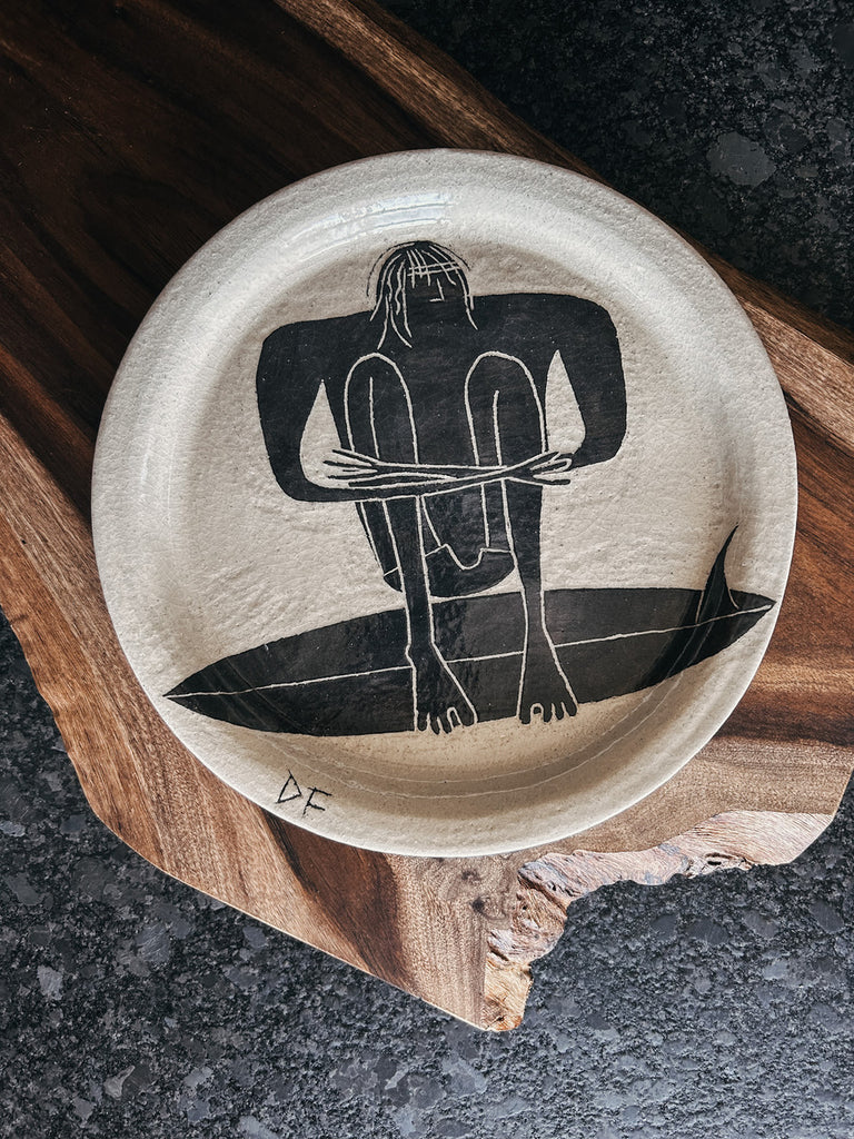 A hand-made ceramic serving platter depicting a man sitting atop his surfboard. Created by Damion Fuller, a ceramicist and surfer based out of Australia.
