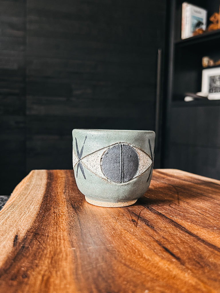 A handcrafted green ceramic cup featuring a sketched eye motif by artist Matthew Ward.