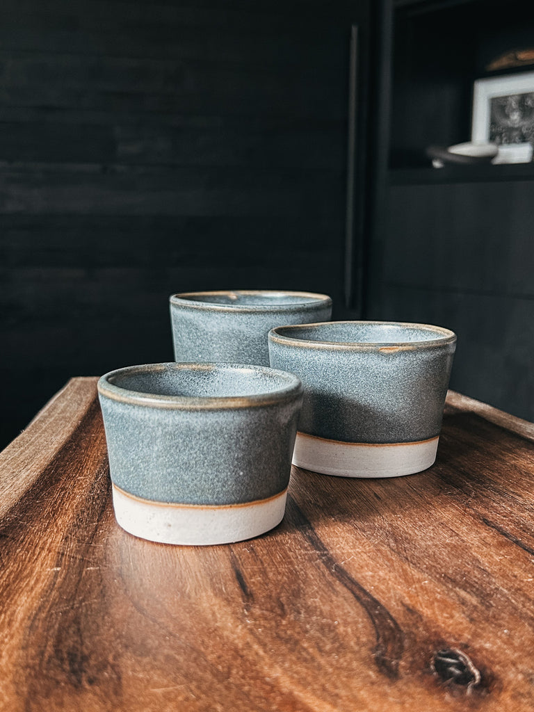 Three ash-glazed, medium-sized ceramic vessels with a soft matte blue-gray finish and raw white clay base. The stackable cups are displayed on a wooden surface, showcasing their minimalist design and handcrafted details.